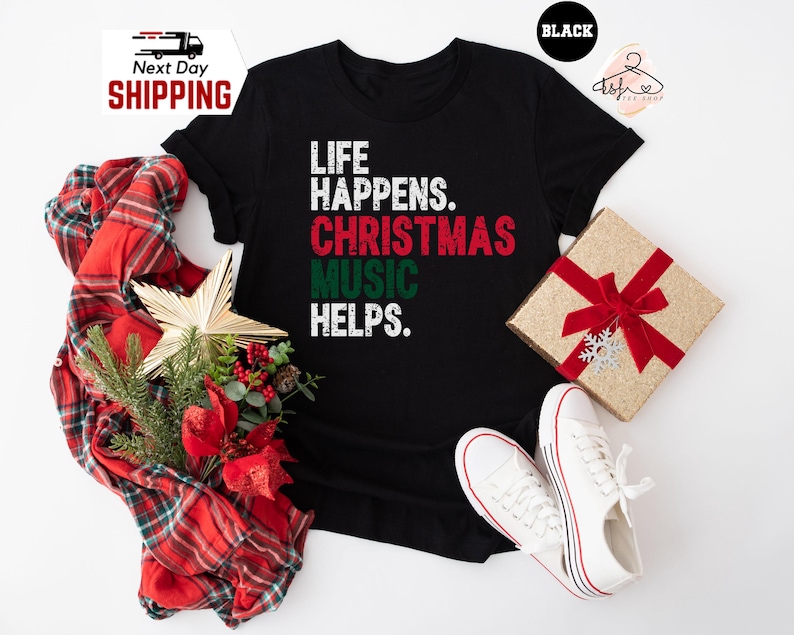 May include: Black t-shirt with the text "LIFE HAPPENS. CHRISTMAS MUSIC HELPS." in white, red, and green. The shirt is displayed with a wrapped gift, white sneakers, and a plaid scarf with Christmas decorations.