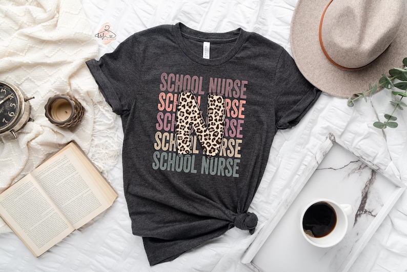 May include: A dark grey t-shirt with the text "SCHOOL NURSE" repeated in a rainbow pattern with a leopard print "N" in the centre.
