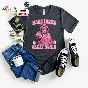 May include: A dark grey t-shirt with a pink bunny rabbit wearing a pink hat and holding an Easter egg. The text "Make Easter Great Again" is printed on the shirt.