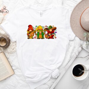 May include: White t-shirt with a graphic of three gnomes wearing sombreros and playing musical instruments. The gnomes are wearing colorful clothing and have festive decorations around them.