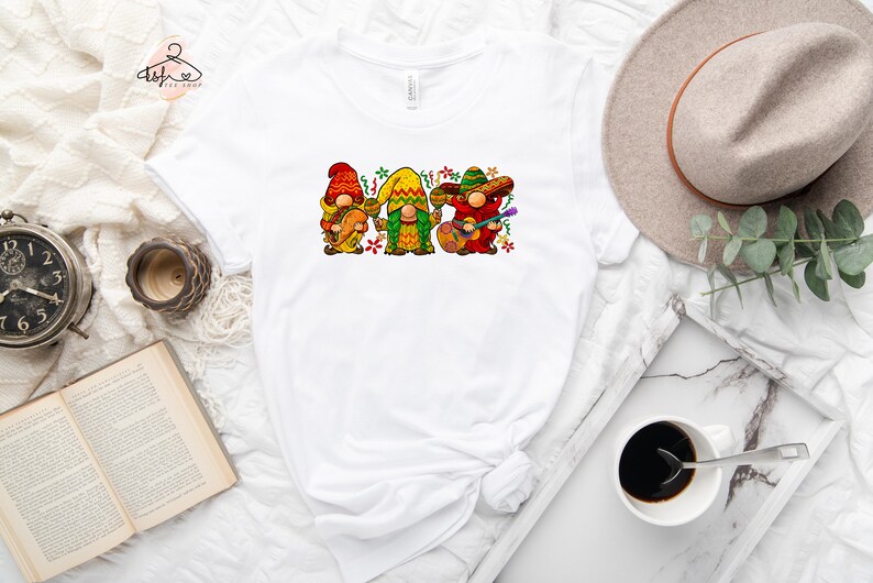May include: White t-shirt with a graphic of three gnomes wearing sombreros and playing musical instruments. The gnomes are wearing colorful clothing and have festive decorations around them.