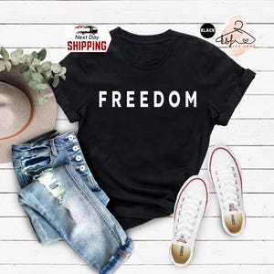 Op de afbeelding: Zwart T-shirt met het woord "FREEDOM" in witte blokletters. Het shirt wordt gecombineerd met een blauwe jeans, witte sneakers en een hoed. De afbeelding staat op een witte houten achtergrond.