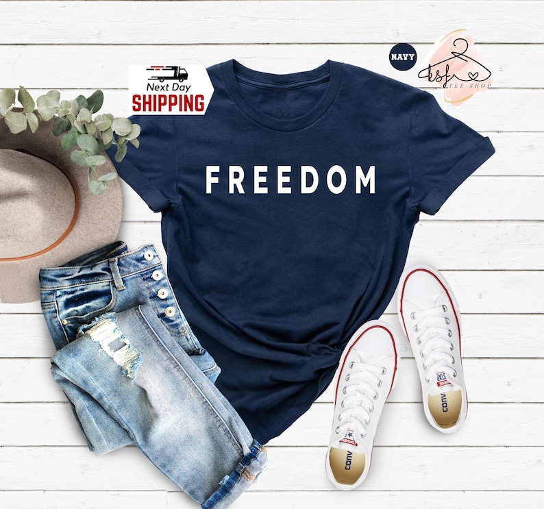 Op de afbeelding: Marineblauw T-shirt met het woord "FREEDOM" in het wit. De afbeelding toont het shirt met lichtblauwe jeans, witte sneakers en een breedgerande hoed. De achtergrond bestaat uit witte houten planken.