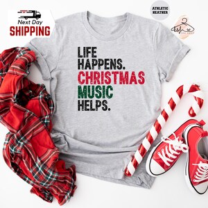 May include: A light gray t-shirt with the text "LIFE HAPPENS. CHRISTMAS MUSIC HELPS." in black, red, and green. The image includes a red and white plaid scarf, a candy cane, and red sneakers.