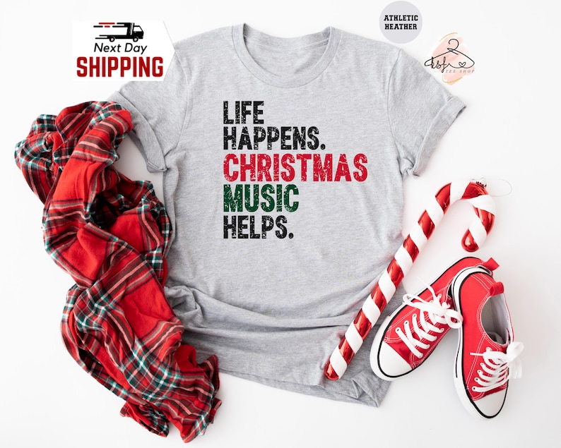 May include: A light gray t-shirt with the text "LIFE HAPPENS. CHRISTMAS MUSIC HELPS." in black, red, and green. The image includes a red and white plaid scarf, a candy cane, and red sneakers.