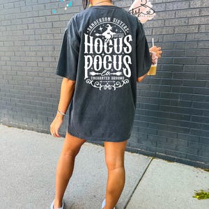 May include: Dark gray oversized t-shirt with the text "Sanderson Sisters Hocus Pocus Co. Enchanted Brooms" in white. The design includes a witch on a broom. The shirt is worn with white sneakers.
