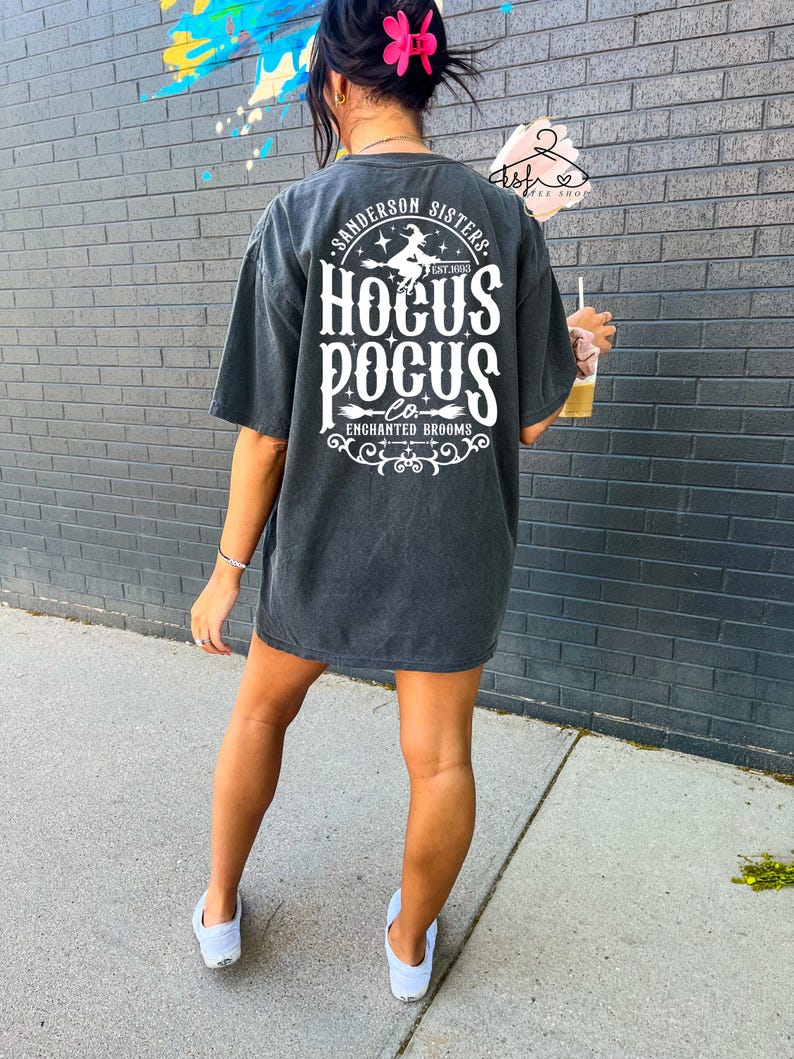 May include: Dark gray oversized t-shirt with the text "Sanderson Sisters Hocus Pocus Co. Enchanted Brooms" in white. The design includes a witch on a broom. The shirt is worn with white sneakers.