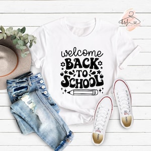 May include: White t-shirt with black text that says "welcome back to school" with a pencil graphic. The shirt is laid out on a white wooden surface with blue jeans and white sneakers.
