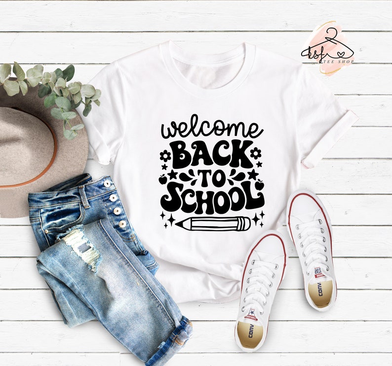 May include: White t-shirt with black text that says "welcome back to school" with a pencil graphic. The shirt is laid out on a white wooden surface with blue jeans and white sneakers.