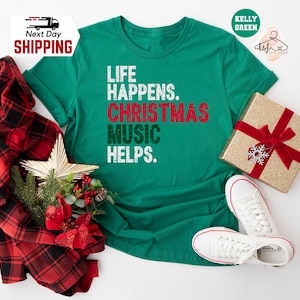 May include: A Kelly Green t-shirt with the text "LIFE HAPPENS. CHRISTMAS MUSIC HELPS." in white and red. The shirt is paired with white sneakers, a wrapped gift box with a red bow, and a red and black plaid scarf. A gold star and Christmas greenery complete the festive scene.