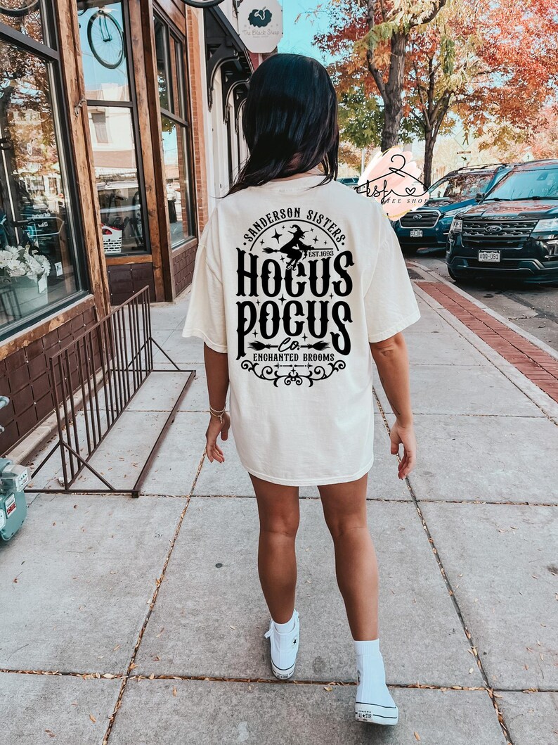 May include: Cream-colored t-shirt with the text "Sanderson Sisters Hocus Pocus Enchanted Brooms" in black. The design includes a witch on a broom. The person is wearing white sneakers and socks.