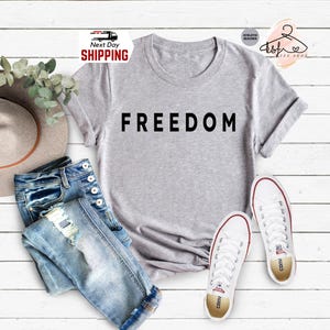 Op de afbeelding: Een lichtgrijs t-shirt met het woord "FREEDOM" in zwarte letters. Het shirt wordt gecombineerd met een blauwe jeans en witte sneakers. Een hoed en groen zijn ook in de afbeelding.