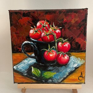 May include: A painting of a still life featuring a black ceramic mug filled with red tomatoes on a vine. The mug is sitting on a blue and white patterned napkin on a wooden surface. The background is a dark red.