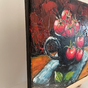 May include: A still life painting of a black mug filled with red tomatoes on a blue and white patterned napkin. The background is a dark red with textured brushstrokes.