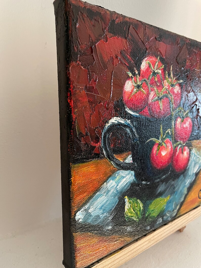 May include: A still life painting of a black mug filled with red tomatoes on a blue and white patterned napkin. The background is a dark red with textured brushstrokes.