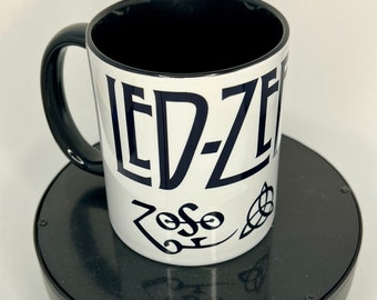 May include: White ceramic mug with black interior and handle. The mug features the band name "LED-ZEP" in bold, dark blue letters, along with the band's logo and the word "Zoso". A classic music-themed gift.