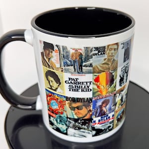 May include: White ceramic mug with a black rim and handle. The mug features a collage of album covers featuring Bob Dylan. The album covers include "Pat Garrett & Billy the Kid", "Shot of Love", "The Times They Are A-Changin'" and "Bob Dylan's Greatest Hits Vol. II".