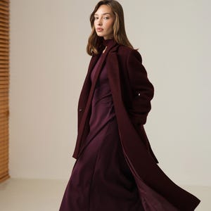 May include: A woman in a long, burgundy coat and dress. The coat is open, showing the dress beneath. The dress has a high neckline and long sleeves. She wears dark burgundy heels. The outfit is a monochromatic style, with a focus on the burgundy colour.