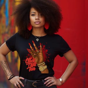 May include: Black t-shirt with a graphic of a hand with long red nails, a gold watch, and the text "Clock That Tea." The shirt is worn with gray jeans and a black belt. The background is a vibrant red.
