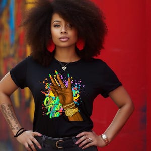 Puede incluir: Camiseta negra con un gráfico colorido de una mano con uñas largas pintadas, un reloj dorado y el texto "Clock That Tea". El diseño incluye salpicaduras de pintura coloridas. La modelo lleva un collar y anillos.