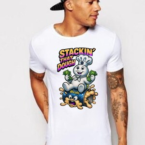 May include: White t-shirt featuring a cartoon image of the Pillsbury Doughboy holding money and sitting on a can of biscuits. The text "Stackin' That Dough" is above the image in a colorful font.