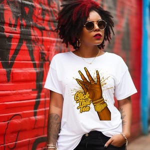 May include: White t-shirt with a graphic of a hand with gold fingernails and jewelry, and the text "Clock That Tea". The shirt is paired with black shorts. The background features a red and black graffiti wall.