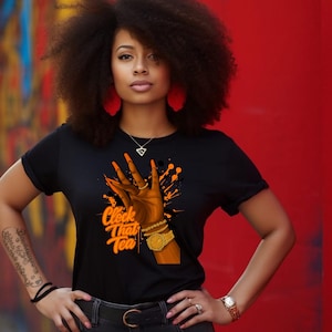 May include: Black t-shirt with the text "Clock That Tea" and a graphic of a hand with orange nails and gold jewelry. The design includes orange paint splatters. The model is wearing a watch and jewelry.