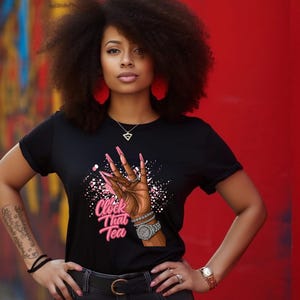 May include: Black t-shirt with a graphic of a hand with long pink nails, a watch, and the text "Clock That Tea." The shirt is worn by a person with dark hair and red earrings, standing in front of a red wall.