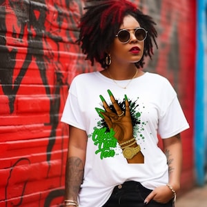 May include: White t-shirt with a graphic of a hand with long green fingernails making a hand gesture. The graphic includes the text "Clock That Tea" in green. The person is wearing a gold watch and bracelets.