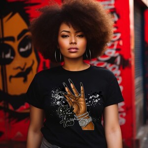 May include: Black t-shirt featuring a graphic of a hand with long black fingernails, a watch, and bracelets. The text "Clock That Tea" is printed below the hand. The background is a red wall with graffiti.