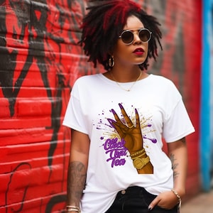 Puede incluir: Camiseta blanca con un gráfico de una mano con uñas largas moradas, un reloj dorado y el texto "Click That Tea" en morado. El diseño está acentuado con salpicaduras doradas y moradas.