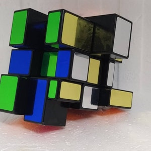 May include: A black puzzle cube with rectangular faces. The cube features green, blue, yellow, and white colored squares. The cube is a twisty puzzle, designed to be solved by rotating the layers.