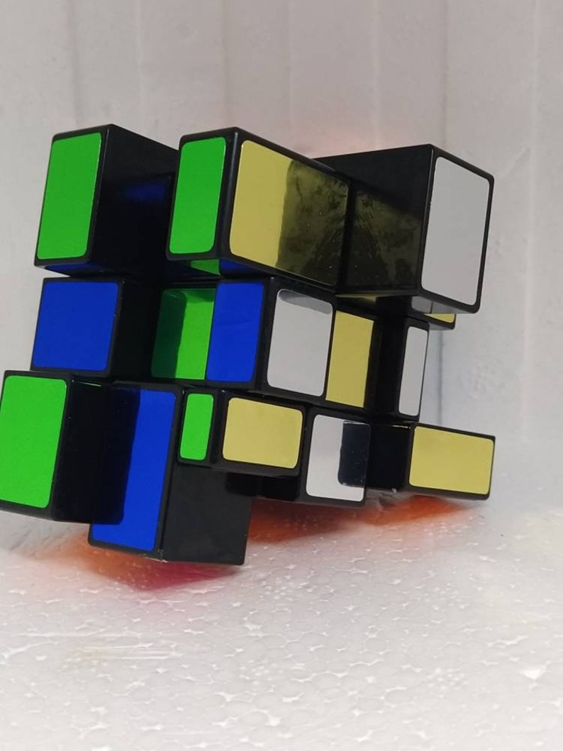 May include: A black puzzle cube with rectangular faces. The cube features green, blue, yellow, and white colored squares. The cube is a twisty puzzle, designed to be solved by rotating the layers.