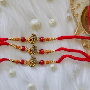 May include: Three red thread bracelets featuring a central gold-toned charm shaped like a peacock feather and the Om symbol. Each bracelet is accented with small red beads, metallic gold spacers, and brown textured beads on either side of the charm.