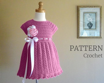 May include: A pink crochet dress for a child, displayed on a mannequin. The dress features a floral embellishment, a white ribbon, and a scalloped hem. The word "PATTERN" and the word "Crochet" are printed on the wall.