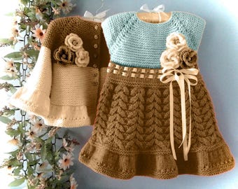 May include: A knitted two-piece outfit for a baby or toddler, comprising a brown and cream cardigan with floral details and a blue and brown dress with a ribbon tie. The dress and cardigan are hanging on a hanger.