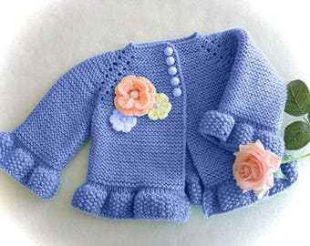 May include: A light blue knitted cardigan with a ruffled hem and sleeves. The cardigan features decorative flowers in peach, yellow, and white. It has five light blue buttons down the front.