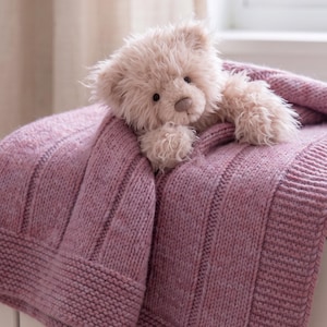 Puede incluir: Un osito de peluche esponjoso de color beige claro con ojos oscuros y nariz marrón, descansando sobre una manta de punto gruesa de color malva. La manta presenta un patrón texturizado con secciones acanaladas verticales y un borde ancho en punto bobo.