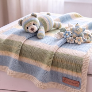 May include: A knitted baby blanket with horizontal stripes in light blue, sage green, and cream. A matching knitted teddy bear and a star-shaped rattle with flower accents rest on top. A brown leather tag reads Knit with Love.