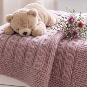 May include: A plush beige teddy bear with a dark brown nose lies on a dusty rose knitted blanket featuring a textured cable knit pattern. A small bouquet of pink and purple flowers with green stems rests beside the bear on the fabric.