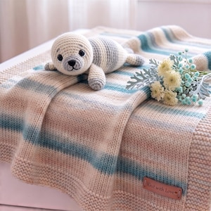 May include: A cream-colored knit blanket with horizontal stripes in shades of blue and beige, featuring a small crocheted seal toy and a brown leather tag with the text Knit with Love. A small bouquet of white and blue flowers rests nearby.