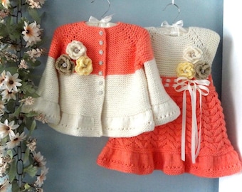 May include: A knitted children's outfit comprising a coral and white cardigan with flower detailing and a matching coral dress. The set is displayed on hangers, each with a white bow.