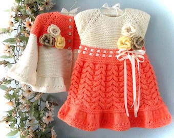 May include: A knitted coral dress and matching cardigan set. The dress has a cream bodice with floral embellishments and a coral skirt with a ruffled hem. The cardigan has floral accents and button closures.