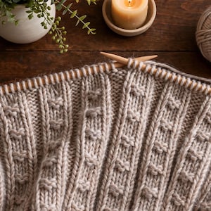 Könnte beinhalten: Ein beigefarbenes Strickstück mit vertikalem Rippenmuster und kleinen, erhabenen knotenartigen Maschen. Zwei helle Holzstricknadeln stecken in der obersten Maschenreihe und halten das dicke, strukturierte Garn fest.