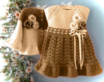 May include: A knitted outfit for a child, comprising a cream-coloured cardigan and a dress. The cardigan is brown and cream with flower embellishments. The dress is cream and brown with a cable knit design and a ribbon bow.