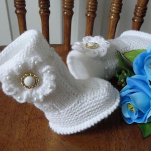 May include: A pair of white knitted baby booties with pearl and gold flower embellishments. The booties are decorated with small pearls around the flower and are sitting on a wooden surface. The booties are next to a bouquet of blue roses.