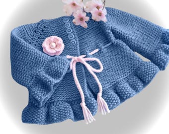 May include: A blue knitted cardigan with a frilled hem and sleeves. The cardigan has a pink flower embellishment and a pink tie closure. Decorative pink flowers are placed on top of the cardigan.