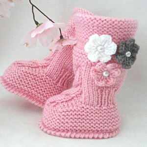 Può includere: Un paio di stivaletti rosa per neonati lavorati a maglia con dettagli a treccia e tre applicazioni floreali decorative. I fiori sono bianchi, grigi e rosa, ciascuno con un centro di perla. Gli stivaletti sono progettati per tenere caldi i piedi del bambino.