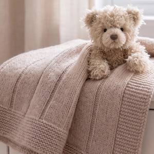 May include: A light beige knitted blanket with a textured vertical ribbed pattern and a wide garter stitch border. A fluffy, light brown teddy bear with dark eyes and a brown nose sits on top of the folded fabric, partially covered by the blanket.