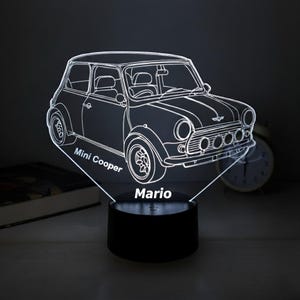 Può includere: Lampada a LED illuminata con il contorno di un'auto Mini Cooper. La lampada presenta il testo "Mini Cooper" e "Mario". La lampada è su una base nera ed emette una luce bianca brillante.